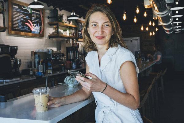 Young woman sitting in cafe drinking coffee using mobile phone.Businesswoman holding cellphone, checking email, planning working project communication online Female working freelance