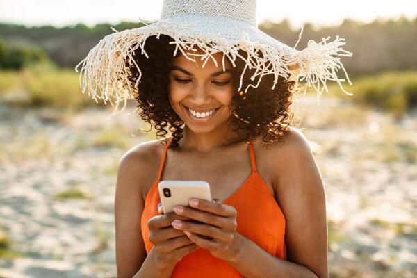 Image of pretty smiling young african woman using mobile phone at the beach outside