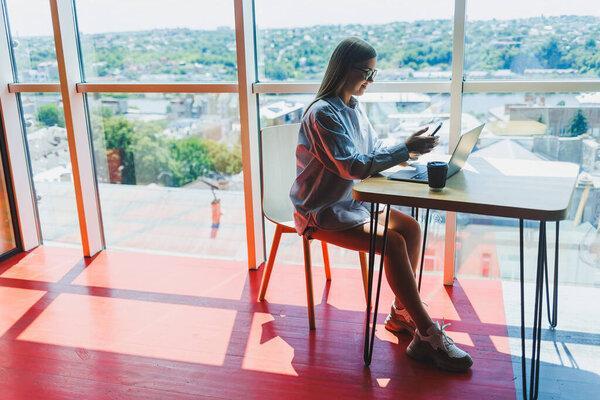 Successful woman is looking at a laptop in a cafe and drinking coffee. A young smiling woman in glasses sits at a table near the window with a phone. Freelance and remote work. Modern female lifestyle