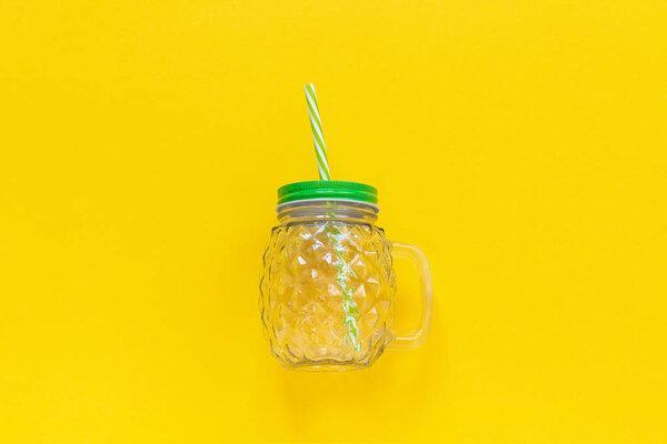 Empty glass jar in form of pineapple with green lid and straw for fruit or vegetable smoothies, cocktails and other beverages on yellow background. Top view Copy space Template