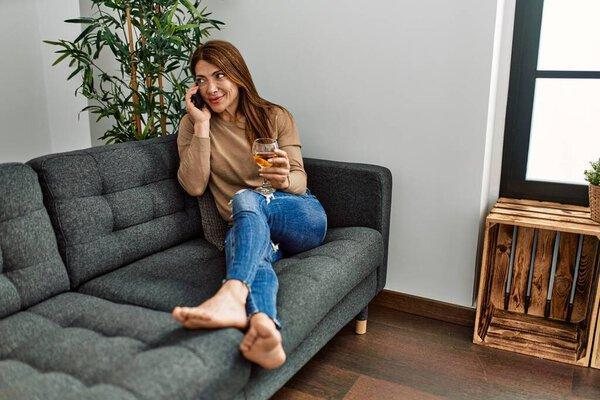 Middle age caucasian woman drinking wine and talking on the smartphone at home.
