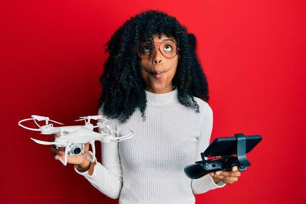 African american woman with afro hair using drone with remote control making fish face with mouth and squinting eyes, crazy and comical. 