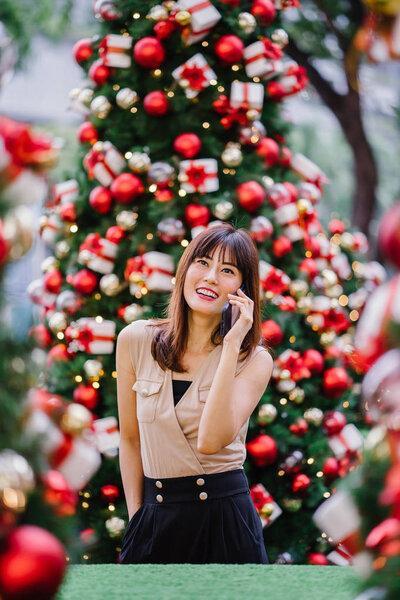 beautiful middle-aged woman is talking happily on her smartphone and smiling as she stands amidst beautifully decorated Christmas trees during the day