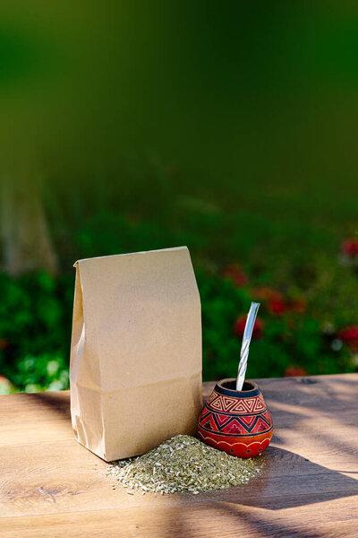 A rustic pack of brown paper with a traditional mate made of calabash over a wooden table with yerba mate scattered over it. Vertical Format.