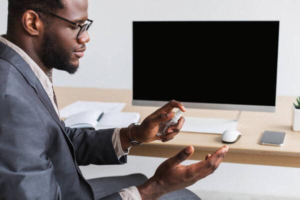 Young black businessman spraying antiseptic on his hands before work at office, mockup for website on pc monitor screen