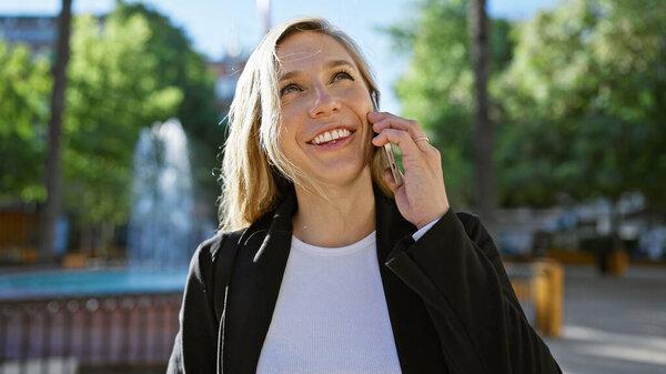 A smiling woman talking on a mobile phone while walking in a sunny urban park with green trees