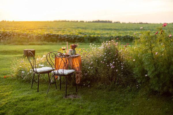Beautiful green lawn with flowers and dining table for two during the sunset. Romantic picnic on nature
