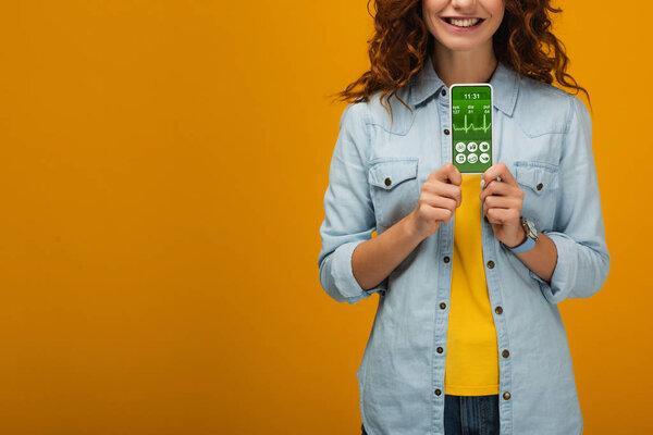 cropped view of cheerful curly woman holding smartphone with e-health app on screen on orange 