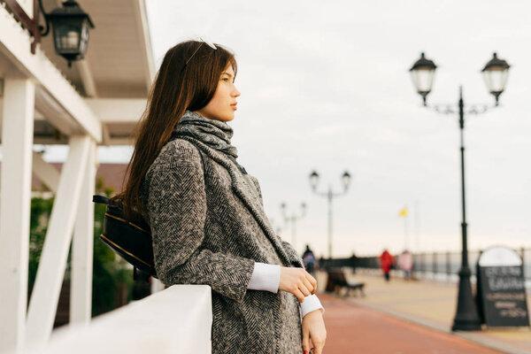 stylish dark-haired young girl in a gray coat walking through the streets of the city, enjoying the overcast weather