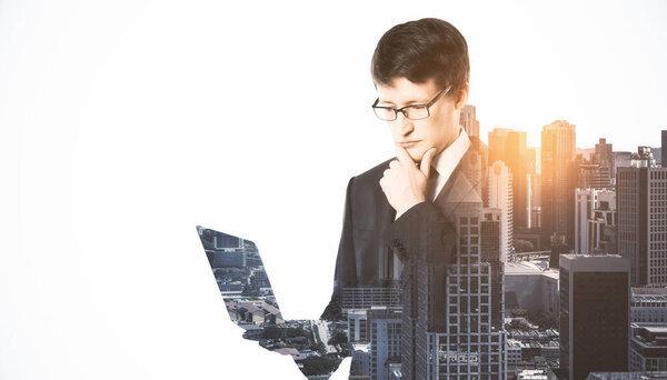 Portrait of attractive thoughtful european businessman using laptop or tablet on bright city background with mock up place. Technology, communication and success concept. Double exposure