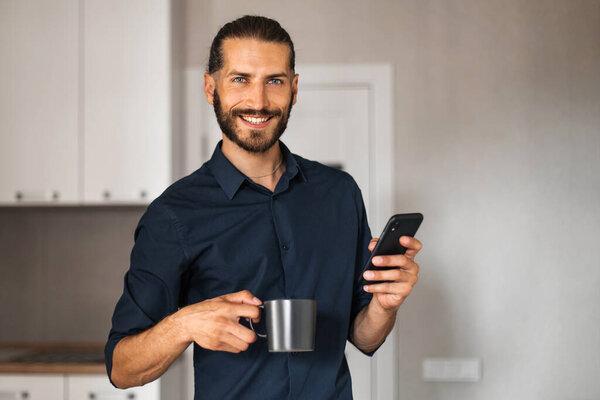 Man smiling, holding a mobile phone and a mug of coffee or tea, looking at the camera