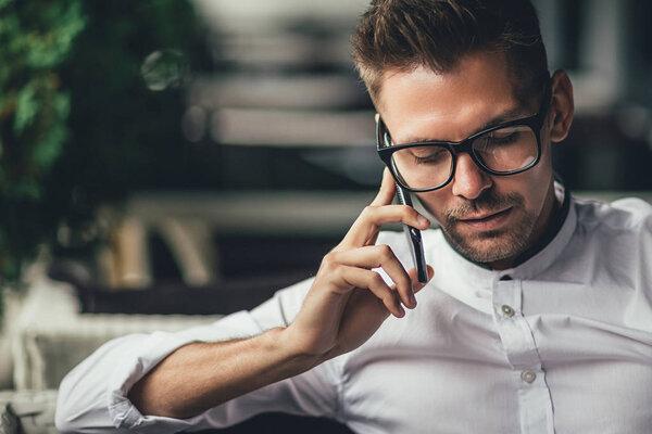 Young handsome businessman is talking on phone, sitting at table in cafe. Man in glasses makes call. Business person in shirt has good