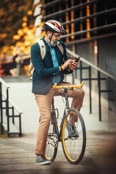 Casual handsome middle-aged businessman is going to the office by bicycle. He is standing on the bike and using smart phone.