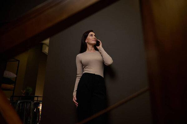 Confident attractive businesswoman, entrepreneur, designer in casual attire talking on mobile phone, gossiping standing on the stairs in the hallway of a modern interior design office. Bottom view