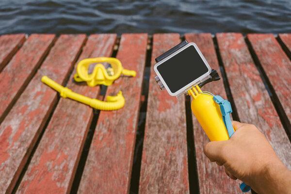 Close-up, action camera mockup with a yellow float in a man's hand. Against the background of a waterproof mask for snorkeling, diving with a tube on a wooden pier with water