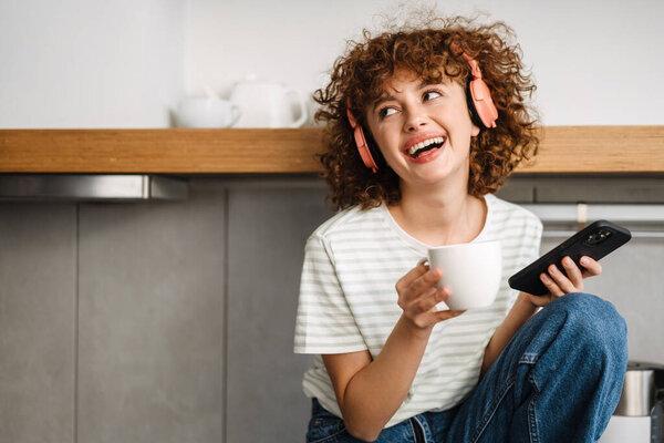 Smiling curly woman in headphones using cellphone while drinking coffee at kitchen