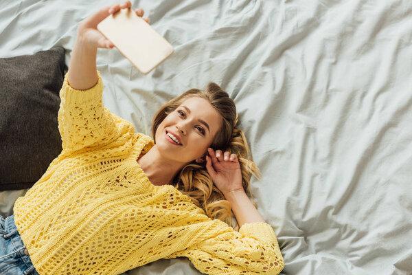 overhead view of beautiful girl lying in bed and taking selfie on smartphone