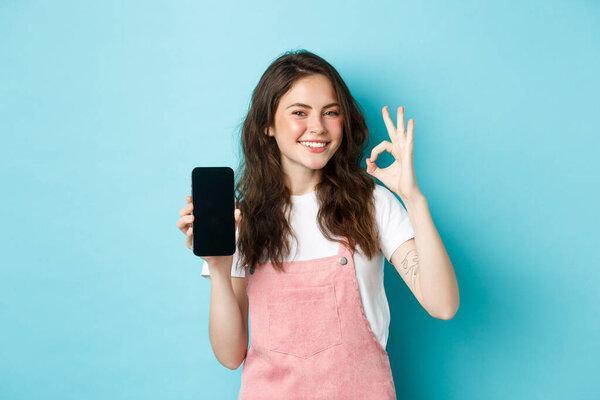 Very good. Smiling young woman showing product online on empty smartphone screen, recommending your brand, make ok sign, standing satisfied against blue background