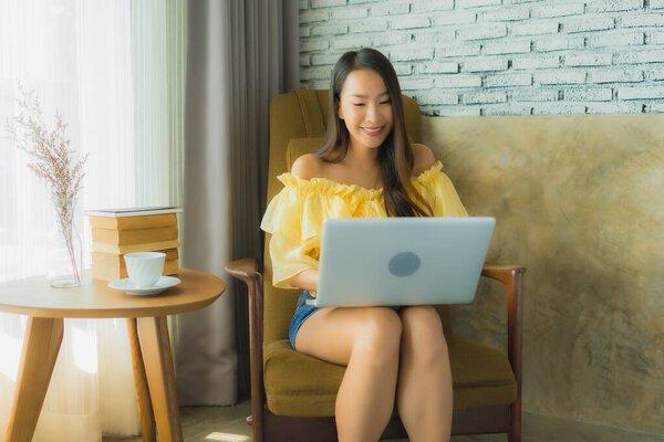 Portrait young asian woman sit on chair using laptop computer with coffee cup and book in living room interior