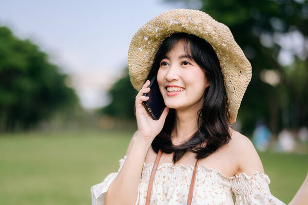 Portrait of young asian woman traveler with weaving hat, basket, mobile phone and camera on green public park background. Journey trip lifestyle, world travel explorer or Asia summer tourism concept.