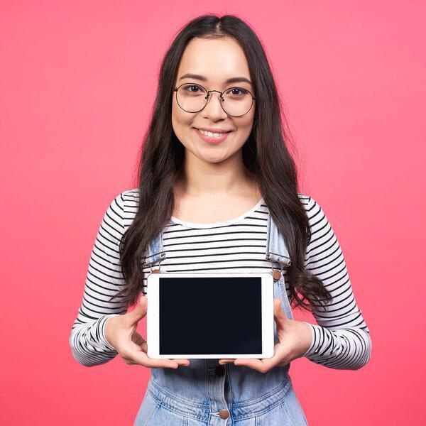 A cute brown-haired young Asian girl, schoolgirl, student, in a striped jacket and round glasses, holds a tablet in her hands, smiles and looks straight.