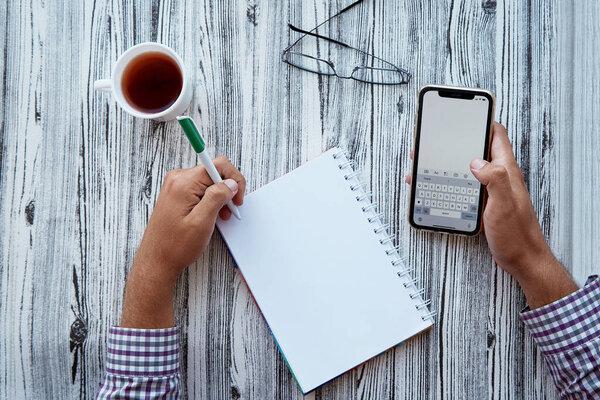 Man writes by his left hand in a notebook while holding a phone. Glasess and cup of tea. Conceptual photo of lefthanded day. Mock up screen phone. Copy space