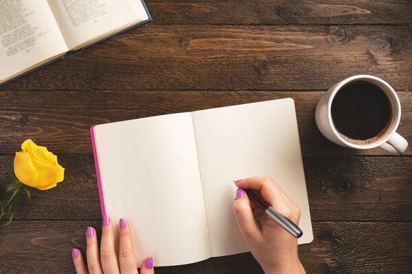 Female hand holding pen. Notebook, cup of coffee, rose, on wooden background. Flat lay, top view, copy space.