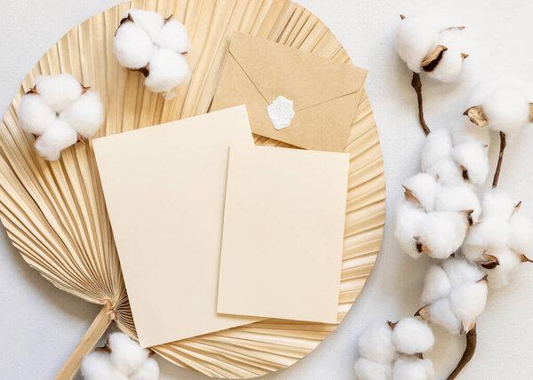 Blank cards and envelope on dried palm leaf with cotton flowers top view, mockup. Romantic scene with vertical paper card. Bohemian pastel greeting or wedding Invitation