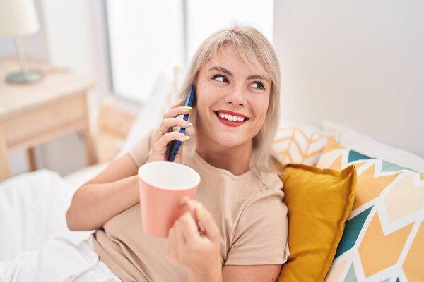 Young blonde woman talking on smartphone drinking coffee at bedroom