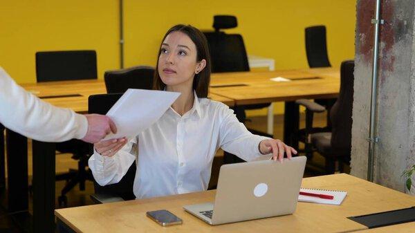 身穿白衬衫和灰色长裤的美丽的白人女商人坐在她的工作台上，从大学拿起文件，阅读，翻阅着书页，面带微笑。慢动作视频.