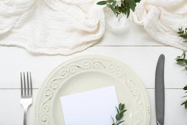 Plate with cutlery, blank card and plant branches on white wooden background