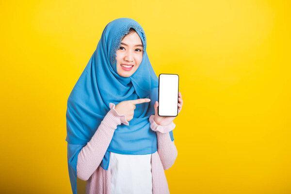 Asian Muslim Arab, Portrait of happy beautiful young woman Islam religious wear veil hijab funny smile she showing blank screen smart mobile phone and point screen isolated on yellow background