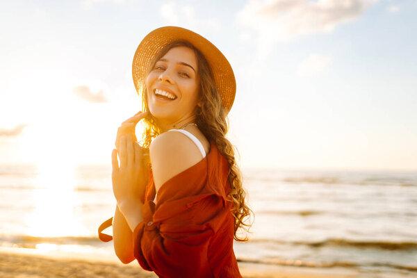 戴帽子的可爱女人在海滩上感到自由和享受日落.穿着时髦衣服的女人在户外呼吸新鲜空气的画像。自由、放松和享受的概念.