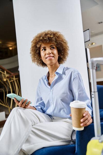 Relaxed and dreamy african american woman with coffee to go and smartphone waiting in hotel lobby