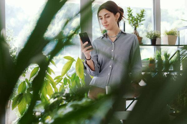 Woman in a room full of plants, she is connecting with her smartphone