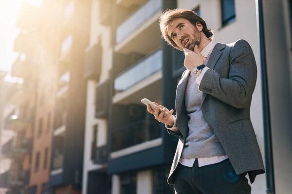 Low angle view of young businessman thinking while text messaging on mobile phone in the city. 