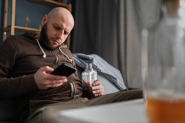 酗酒和人的概念。喝醉了的男人带着智能手机和威士忌坐在沙发上在家里。复制空间