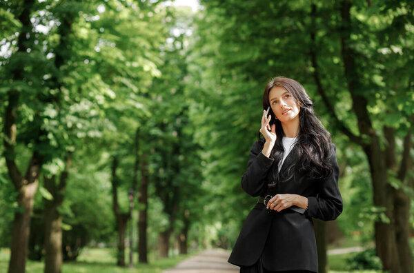 Business woman use cellphone outdoors. Beautiful focused young girl wearing a formal suit in the park.