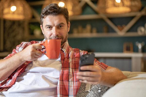 Man looking at phone at home, browsing mobile apps, reading news, chatting or shopping online, holding smartphone. Man in casual clothes with cup coffe at the living room, talking on mobile phone