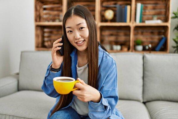 Young chinese girl talking on the smartphone and drinking coffee sitting on the sofa at home.