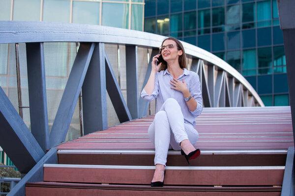 Happy smiling business woman talking on mobile smartphone in the street with office buildings in the background.