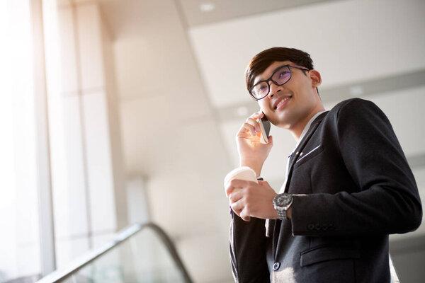 A asian handsome young businessman holding his hot coffee cup while using his phone and standing on the escalator. Business concept.