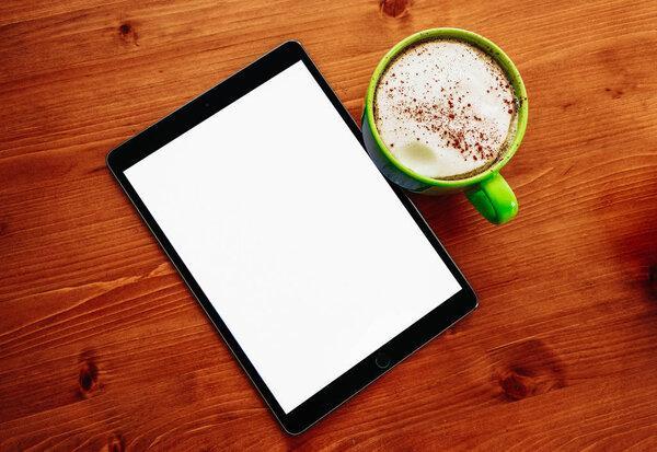 A big green cup of cappuccino coffee on the office wooden table, and an ipad  tablet mockup with blank screen.