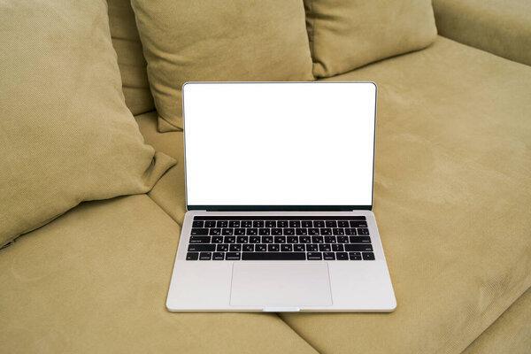 A gray laptop with a white blank screen on a green sofa in the room. The concept of remote work and training, advertising of online products and courses. Mockup for your design