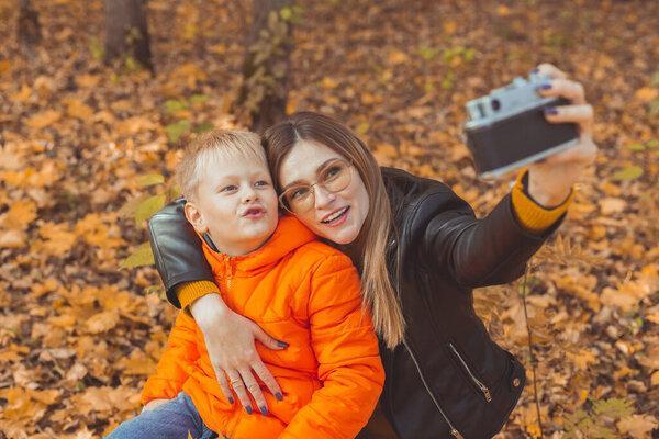Son and mother are taking selfie on camera in autumn park. Single parent, leisure and fall season concept.