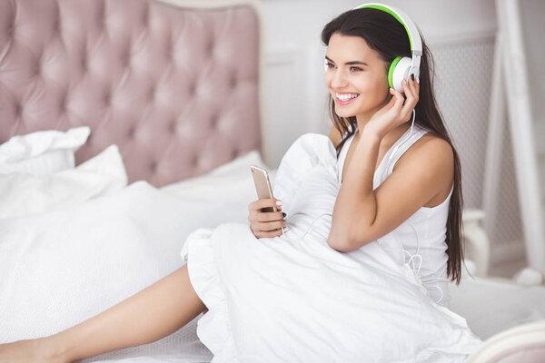 Young attractive woman listenning to the music. Attractive girl in the headphones. Woman indoors in the bed relaxing. Lady wearing earphones.