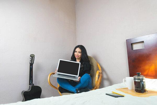 beautiful latin woman showing laptop screen, sitting on sofa in her room
