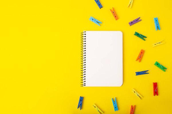 Top view of blank notebook on yellow background with multicolored decorative clothespins around.