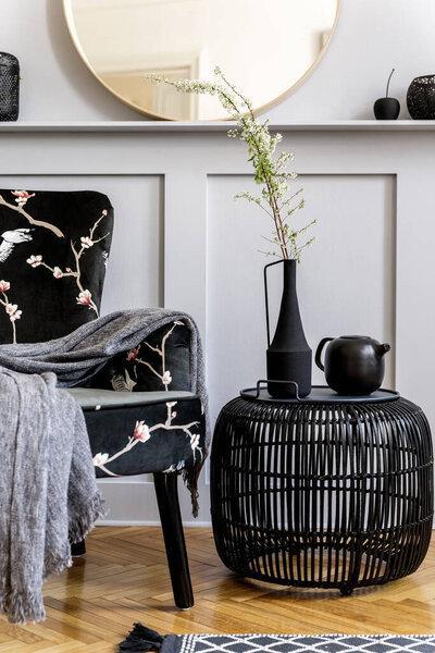 Modern concept of home staging with design armchair, round mirror, black rattan coffee table, leaves in vase, carpet, decoration and elegant personal accessories at stylish living room.