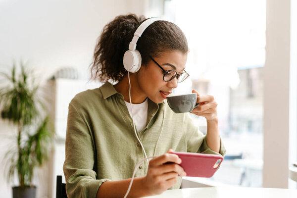 Black woman drinking coffee while using cellphone and headphones in cafe indoors